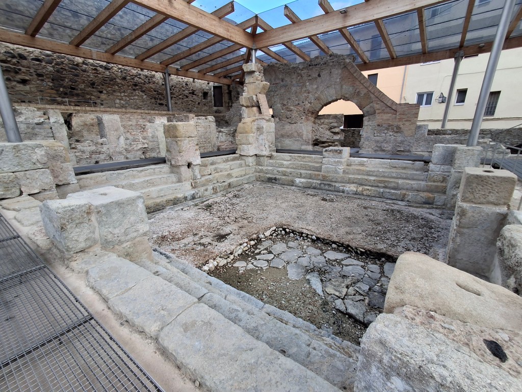 Ruins of an ancient structure with stone walls, partially covered by a transparent roof, showcasing steps and a central space with exposed ground.