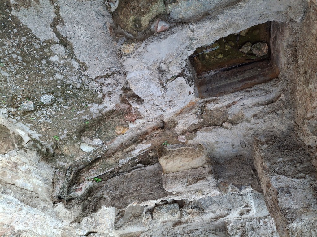 View of a weathered stone ceiling with a small rectangular opening and patches of greenery.