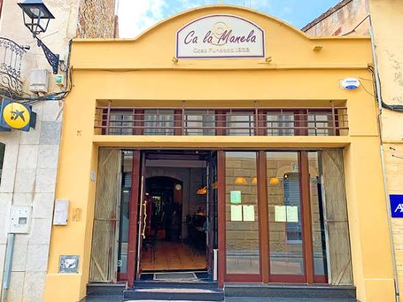 Front view of a restaurant named 'Ca la Manela,' featuring a yellow facade and large glass doors, with menus displayed on the windows.